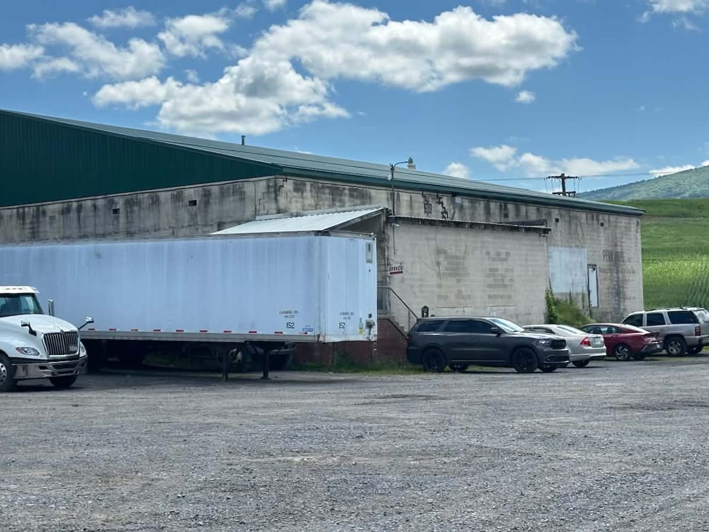 The Penn Turf building in Hollidaysburg, with its green roof and painted signage, trucks and a loading trailer in the yard, and farmland on the ridge behind